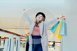 Happy, excited young fashion woman with raised hands holding paper shopping bags on mall background. Woman enjoying new