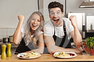 Happy excited young friends loving couple chefs on the kitchen eat tasty pasta make winner gesture.