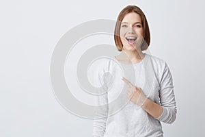 Happy excited young woman in longsleeve with opened mouth feels confident and pointing to the side isolated over white background