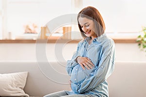 Happy expectant pregnant young woman sits on living room sofa