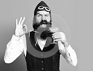 Happy face holding glass of alcoholic beverage in vintage suede leather waistcoat with hat and glasses on red studio