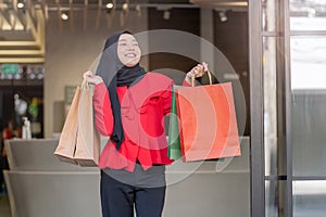 Happy face muslim woman in red dress holding shopping bag and walking out shop building. Concept people activity in shopping