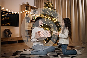 Happy family, attractive young mother and preschool little daughter girl sitting with gift near tree