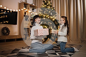 Happy family, attractive young mother and preschool little daughter girl sitting with gift near tree