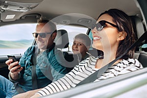 Happy family auto traveling concept image. Car interior view of female driving, man dealing mobile phone and little son smiling photo