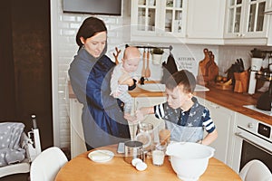 Happy family baking together in modern white kitchen. Mother, son and baby daughter cooking in cozy weekend morning