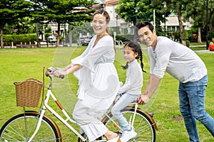 Happy family with bicycle outdoors on summer day, Asian father and mother with daughter enjoying riding a bike outdoors in a park