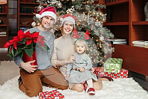 Happy family celebrating Christmas or New Year winter holiday at home. Smiling Caucasian mother and father with baby girl sitting