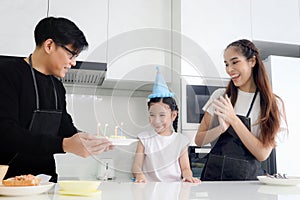 Happy family celebrating daughter girl birthday at kitchen together, father holding cake with candles for child kid to blow,