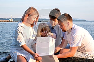 Happy family celebrating little girl's birthday on the seashore mom brings a box with cake girl opens a gift dad mom