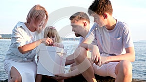 Happy family celebrating little girl's birthday on the seashore mom brings a box with cake girl opens a gift dad mom