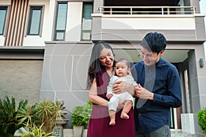 happy family, cheerful father and mother holding and playing with newborn baby while standing in front of their house