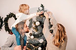 Happy family with child decorate christmas tree. Little girl sitting on daddy`s shoulders hangs ball on the Christmas tree.