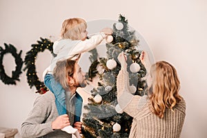 Happy family with child decorate christmas tree. Little girl sitting on daddy`s shoulders hangs ball on the Christmas tree.