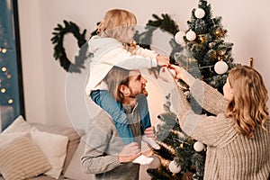 Happy family with child decorate christmas tree. Little girl sitting on daddy`s shoulders hangs ball on the Christmas tree.