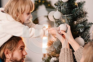 Happy family with child decorate christmas tree. Little girl sitting on daddy`s shoulders hangs ball on the Christmas tree.