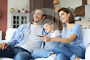 Happy family with child sitting on sofa watching tv, young parents embracing daughter relaxing on couch together