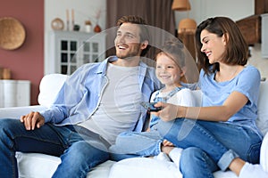 Happy family with child sitting on sofa watching tv, young parents embracing daughter relaxing on couch together