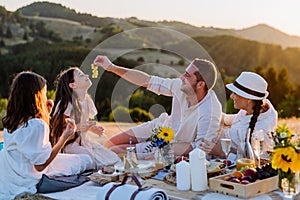 Happy family with children having picnic in park, parents with kids sitting eating healthy meals outdoors.