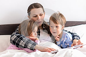 happy family. Close up loving mother lying with daughter son two kids pajamas in bed children reading interesting
