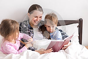 happy family. Close up loving mother lying with daughter son two kids pajamas in bed children reading interesting