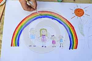 Happy family concept. Child draws on a sheet of paper: father, mother, boy and girl hold hands against background of rainbow and