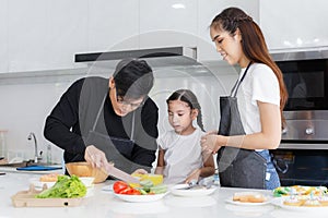 Happy family cooking together in the kitchen. Father, mother, and cute little daughter turn vegetables to make salads