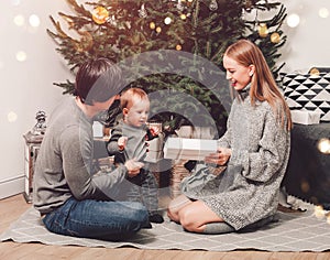 Happy family couple give gifts in the living room, behind the decorated xmas tree, the light give a cozy atmosphere. New Year