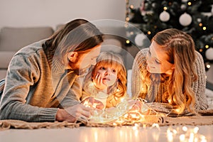 Happy family with daughter lies on the floor and cozy talks by the light of garlands against the background of Christmas tree.