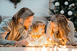 Happy family with daughter lies on the floor and cozy talks by the light of garlands against the background of Christmas tree.