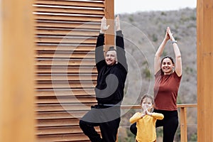 Happy family doing yoga outdoors. Attractive mom, handsome dad and cute daughter are standing in tree pose