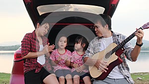 Happy family enjoying road trip on summer vacation. Mother and child sit in the trunk of the car singing along with dad playing th