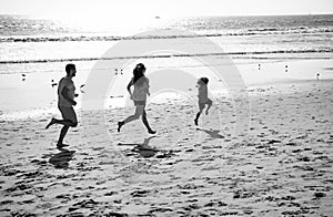 Happy family father, mother and baby son jogging outdoors together, child run by water pool along sea surf on black sand