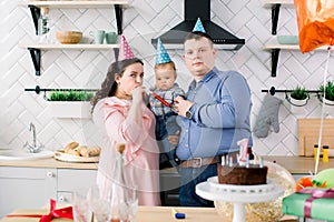 Happy Family, father, mother and little baby boy in party hats blowing in party horns on birthday party at home
