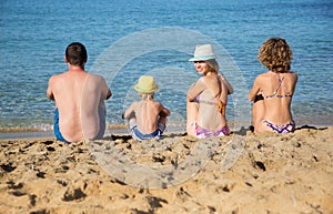 happy family of four people sit with their backs on sandy beach against backdrop of blue sea