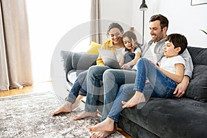 Happy family of four using laptop sitting on the sofa in cozy apartment, parents and two kids talking with relatives