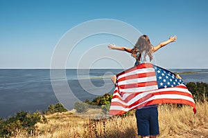 A happy family. Girl sitting on her father& x27;s shoulders with American flag on the shore of the bay. Freedom concept