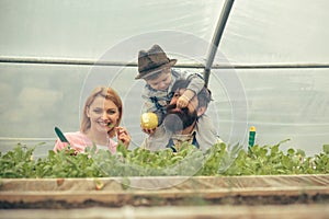 Happy family in greenhouse. Mum posing with green leaf in her mouth while kid is feeding dad with apple sitted on his