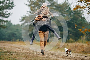 Man carries his wife and daughter on his back at forest path next to dog during walk