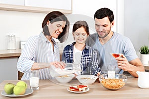 Happy family have healthy breakfast together. Smiling mother pours milk in bowl with cornflakes, eat apples, snacks and