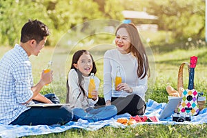 Happy family having fun outdoor sitting on picnic blanket drinking orange juice from glass bottle