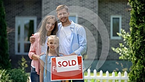 Happy family holding sold sign, standing against new bought house, thumbs up