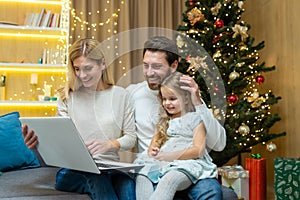 Happy family home for Christmas, husband wife and little daughter sitting on sofa in living room near Christmas tree