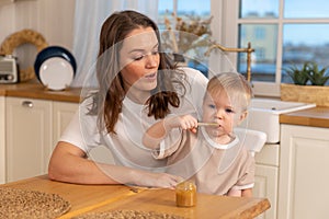 Happy family at home. Mother feeding baby in kitchen. Little boy with messy funny face eats healthy food. Child learns