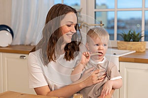 Happy family at home. Mother feeding baby in kitchen. Little boy with messy funny face eats healthy food. Child learns