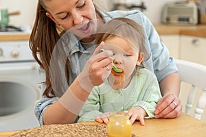 Happy family at home. Mother feeding her baby girl from spoon in kitchen. Little toddler child with messy funny face