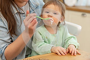 Happy family at home. Mother feeding her baby girl from spoon in kitchen. Little toddler child with messy funny face