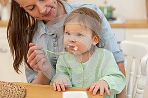 Happy family at home. Mother feeding her baby girl from spoon in kitchen. Little toddler child with messy funny face