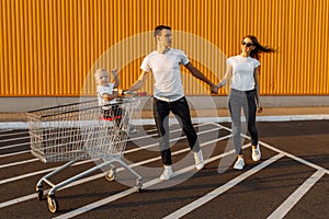 Happy family, husband and wife with a child have fun and run around with a shopping cart, against the background of a yellow wall