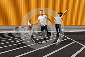 Happy family, husband and wife with a child have fun and run around with a shopping cart, against the background of a yellow wall